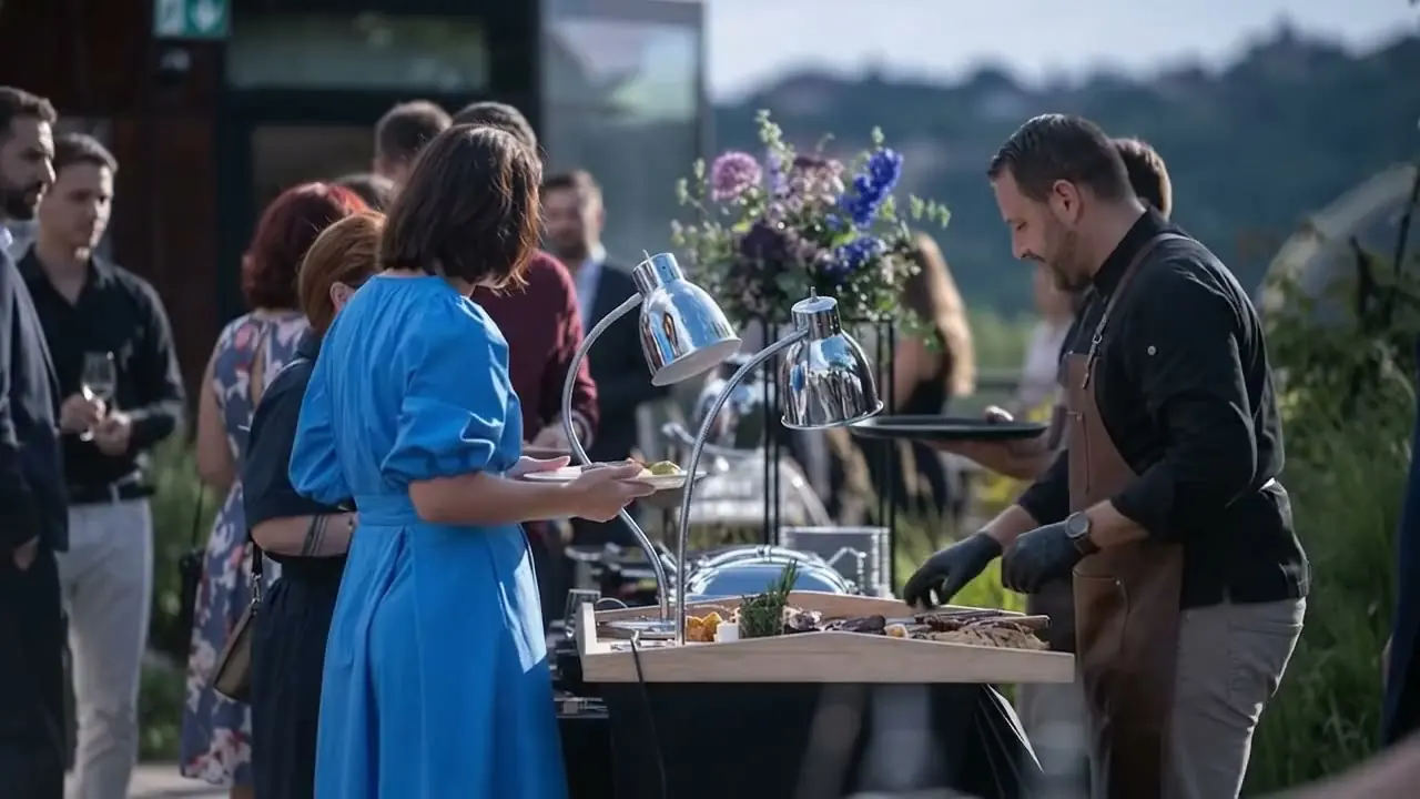 Honza carving steaks under heat lamps on a rooftop above Prague while guests wait with plates