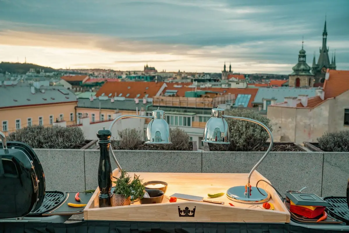 Two Weber Traveler grills and a carving board under heat lamps on a rooftop overlooking Prague’s spires